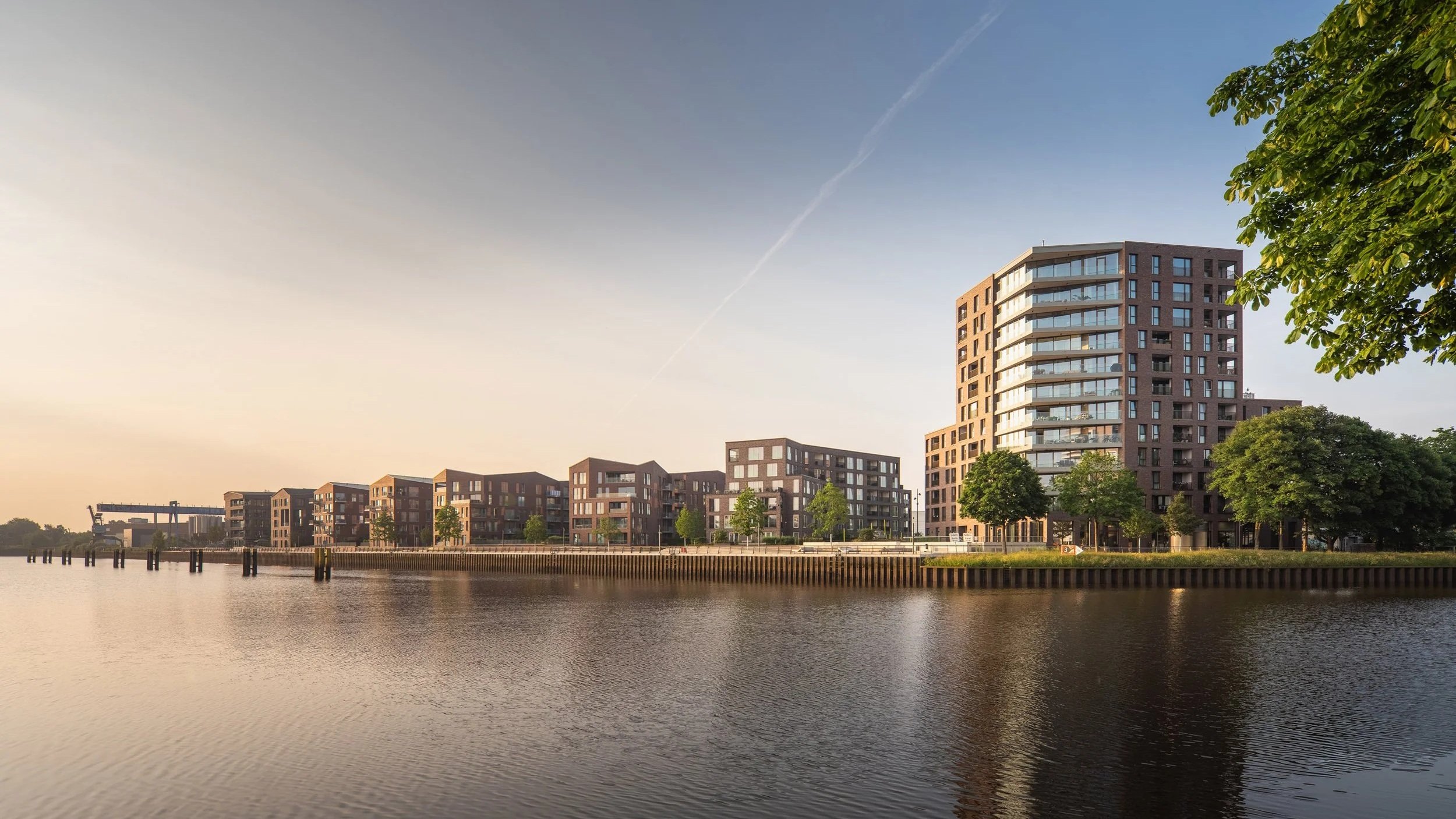 Moderne Wohngebäude am Wasser bei Sonnenaufgang oder Sonnenuntergang, mit Bäumen im Vordergrund.