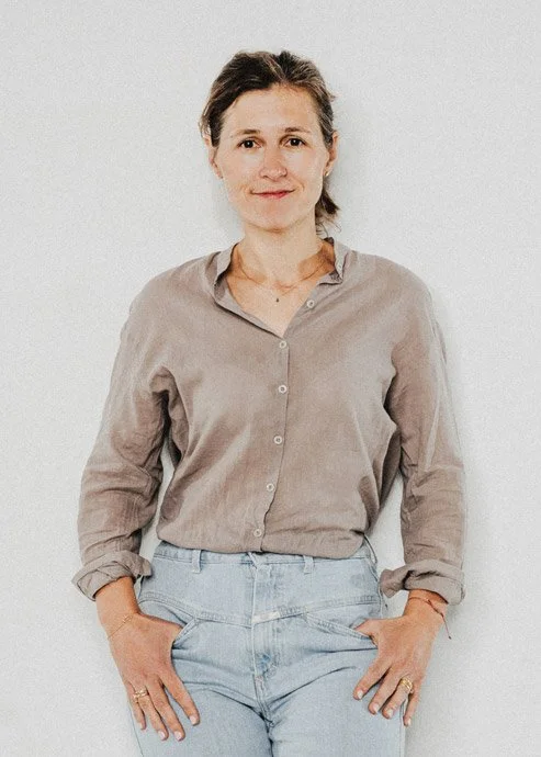 Eine Frau steht vor einem weißen Hintergrund, trägt eine beige Bluse und hellblaue Jeans, lächelt leicht.