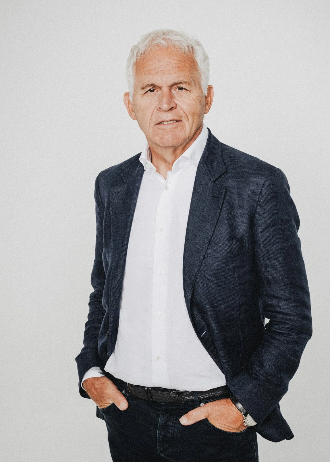 Ein älterer Mann mit weißen Haaren trägt einen dunklen Anzug und ein weißes Hemd, steht vor einfachem hellgrauem Hintergrund, mit einer Hand in der Tasche.