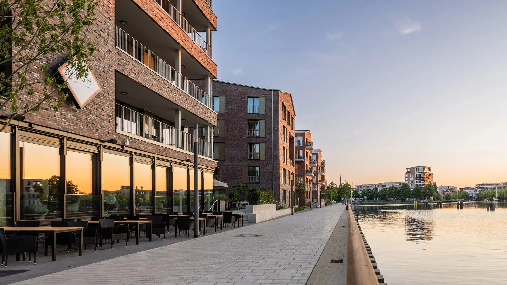 Moderne Gebäude am Wasser bei Sonnenuntergang, mit Balkon und Terrasse, reflektierende Fenster, ruhiger Fluss, Bäume und Himmel im Hintergrund.