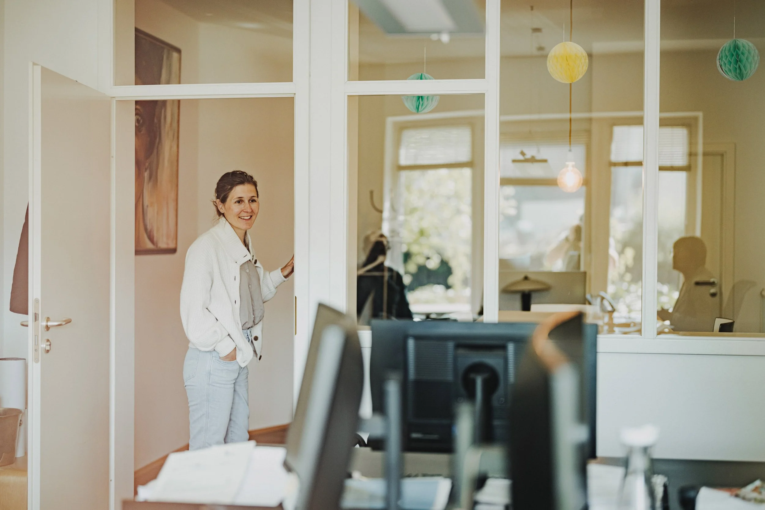 Eine lächelnde junge Frau steht in einem hellen Büro, schaut durch eine Fensterwand. Im Hintergrund sind weitere Personen in einem Raum zu sehen, mit bunten Papierlaternen an der Decke.