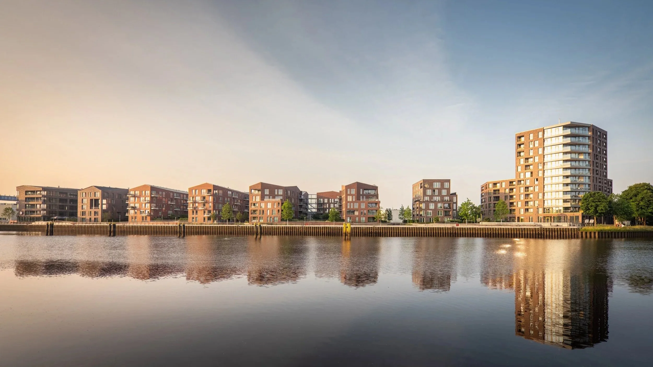Moderne Wohngebäude am Wasser mit spiegelnder Wasseroberfläche bei Sonnenaufgang oder Sonnenuntergang.