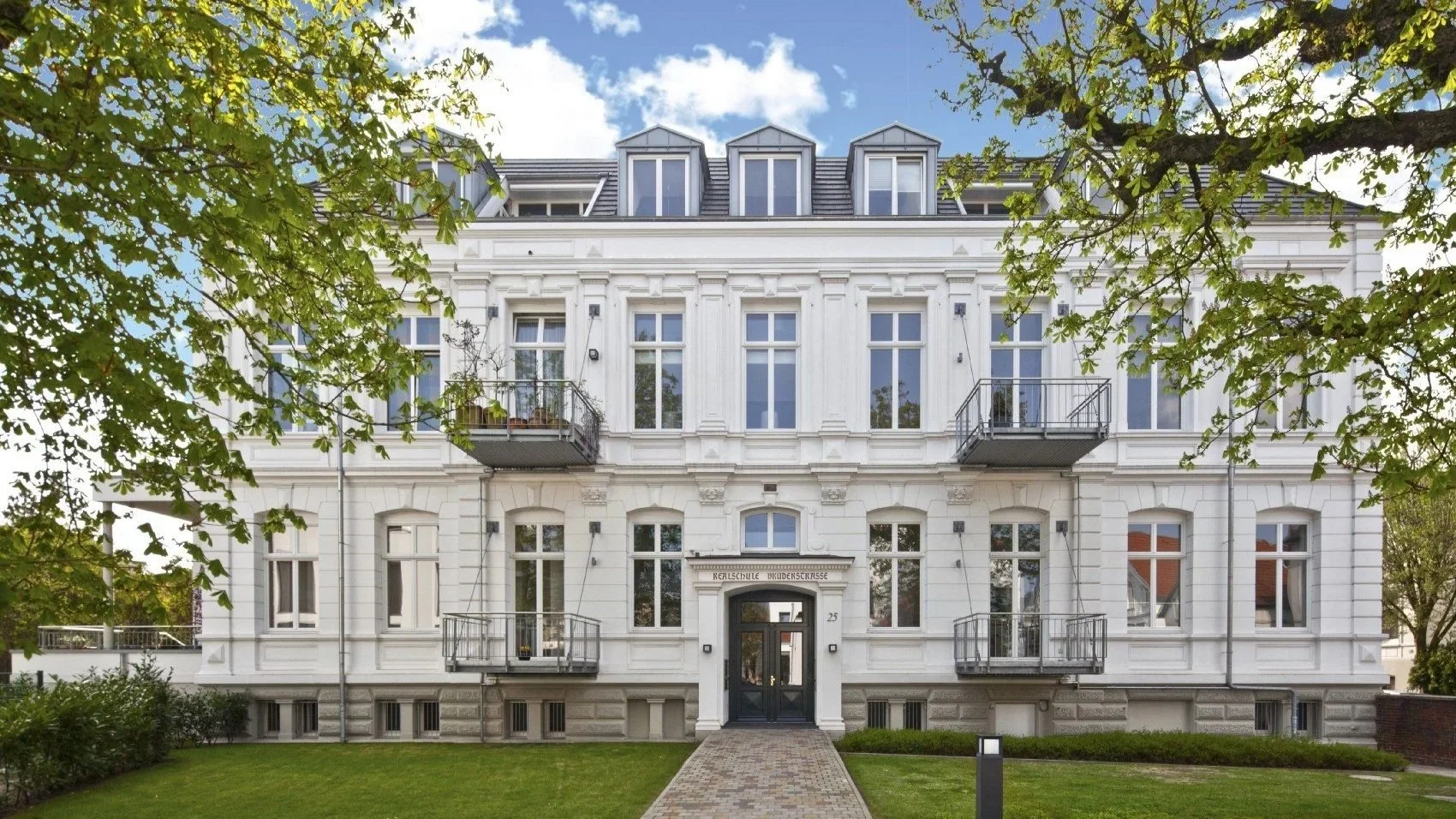 Helles, mehrstöckiges historisches Gebäude mit Balkonen und Garten, erkennbar als eine Bildungseinrichtung mit Schild 'Kreisrealschule Brudernstraße' über dem Eingang.