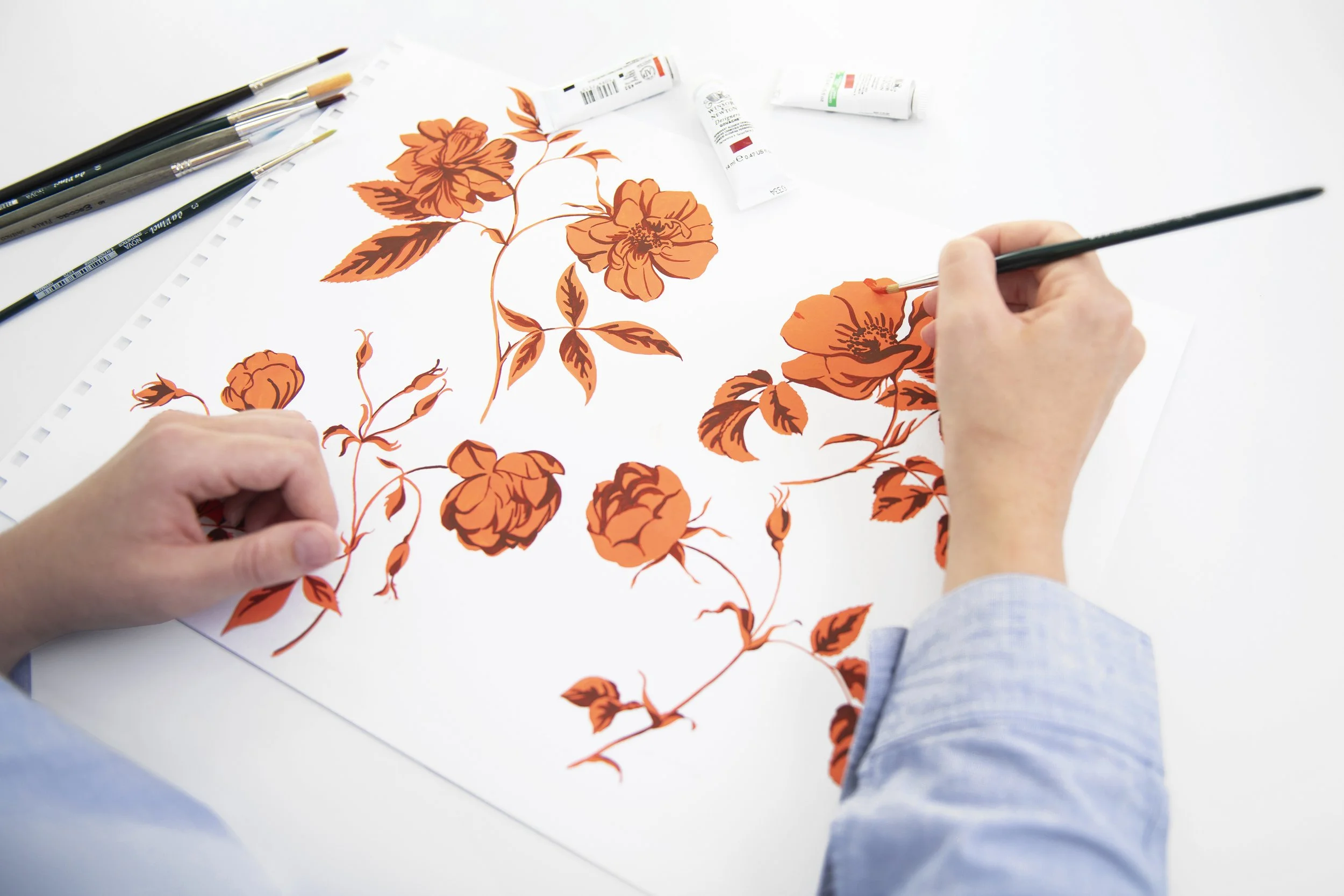 An artist paints orange floral patterns on a white canvas using small brushes, with tubes of paint and additional brushes nearby.