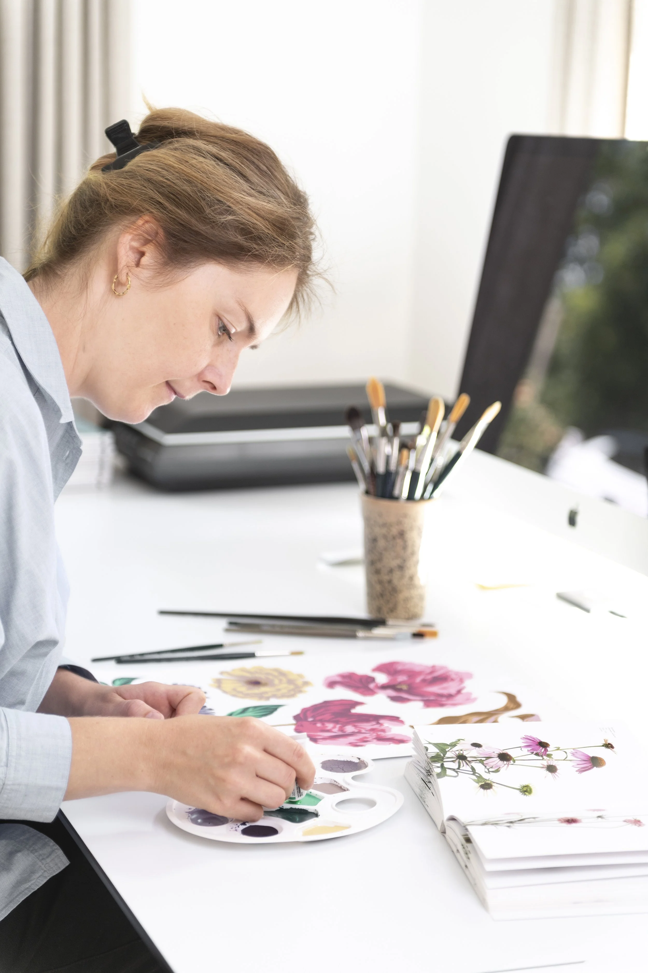 A woman is painting floral illustrations at a white desk. There are paints, brushes, and flower pictures on the desk, with a computer and a container of brushes in the background.