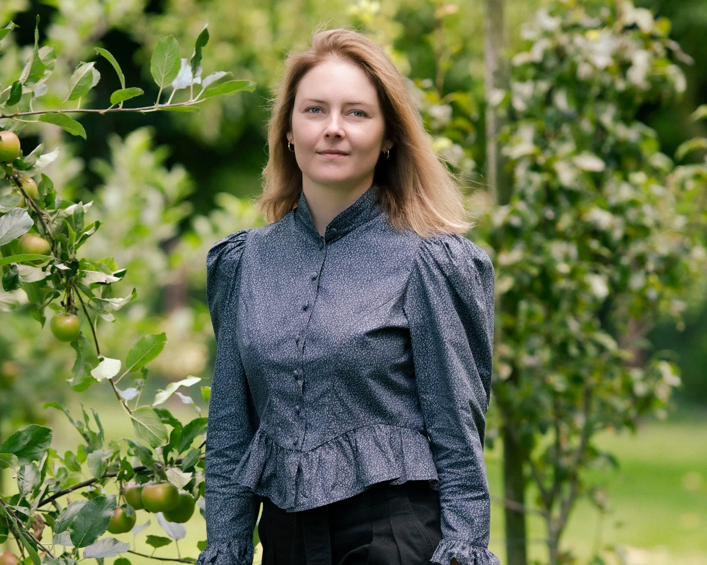 A woman standing outdoors among apple trees, wearing a dark blue long-sleeve blouse with a peplum, in a green orchard setting.