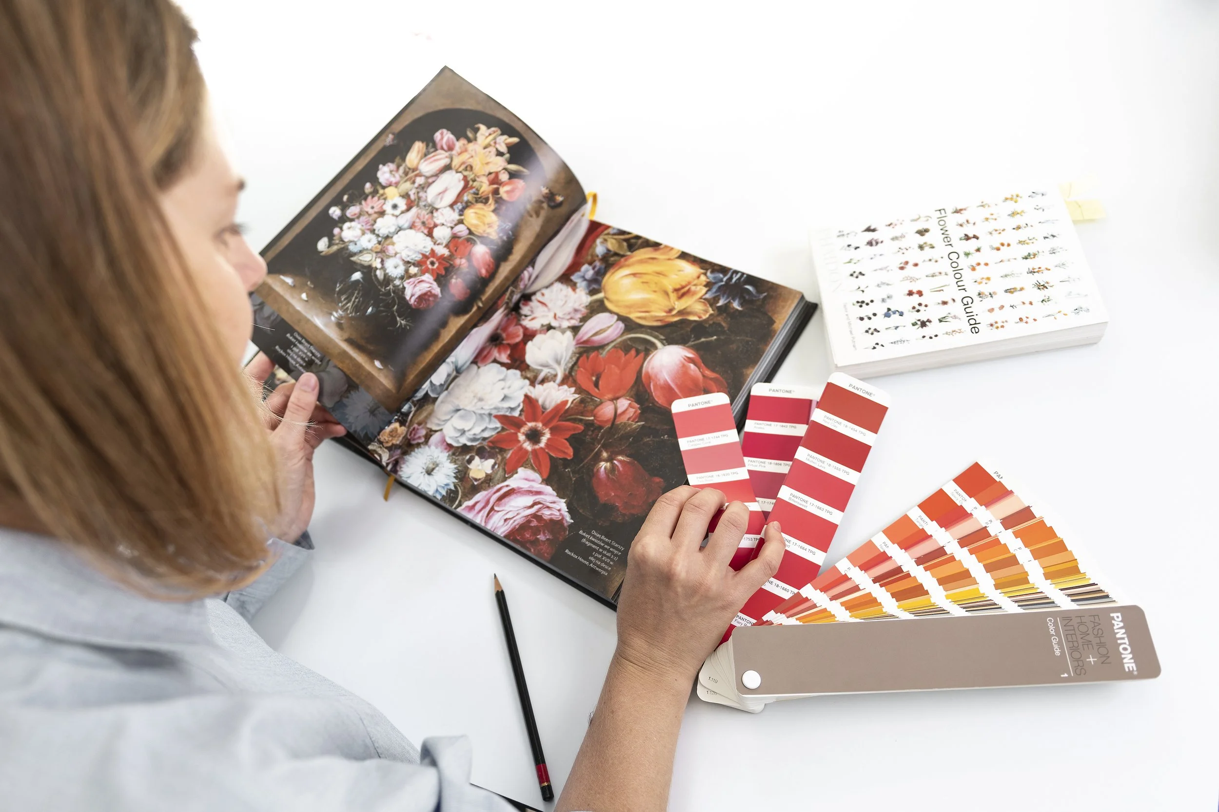 Woman studying color swatches and flower images at a desk with flower photographs, a flower color guide, and an open book with floral images.