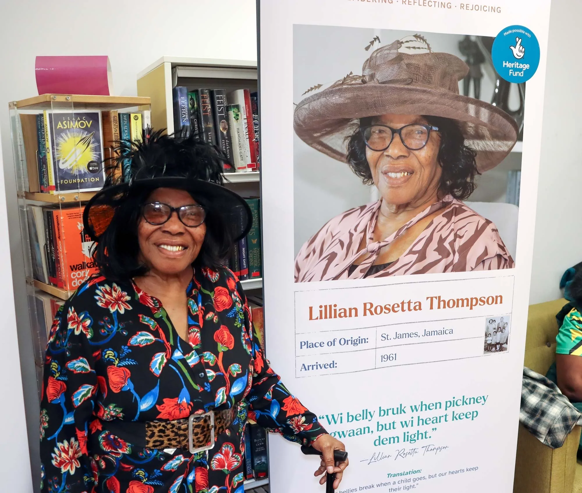 Smiling elderly woman in a black feathered church hat sat smiling next to a roller banner featuring her headshot