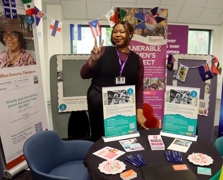Young black woman at an event stand with a Walsall Windrush Sisters oral story banner, table of pens and sweets and bunting.