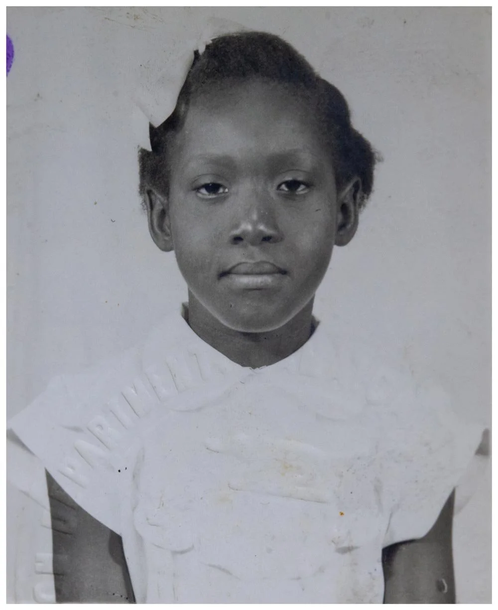 A young Nerissa Harvey photographed as a child in a ruffled white dress, looking solemnly at the camera — an early portrait preserved within the Walsall Windrush Sisters Caribbean heritage archive.