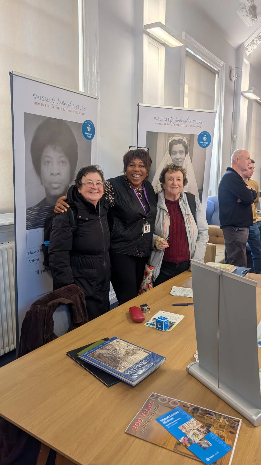 Three women smiling happily with arms on each other's shoulders standing in front of two Walsall Windrush Sister Oral Story Pull-Up Banners
