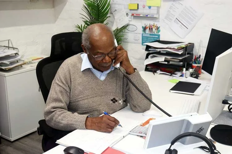 Elderly black man writing into a open set of folders whilst holding a landline handset to his ear