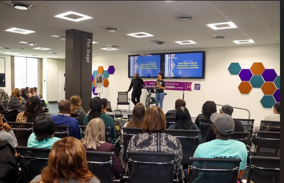 A seated crowd of people facing a black man and young black woman holding a microphone on a stage in front of two wall-mounted screens.