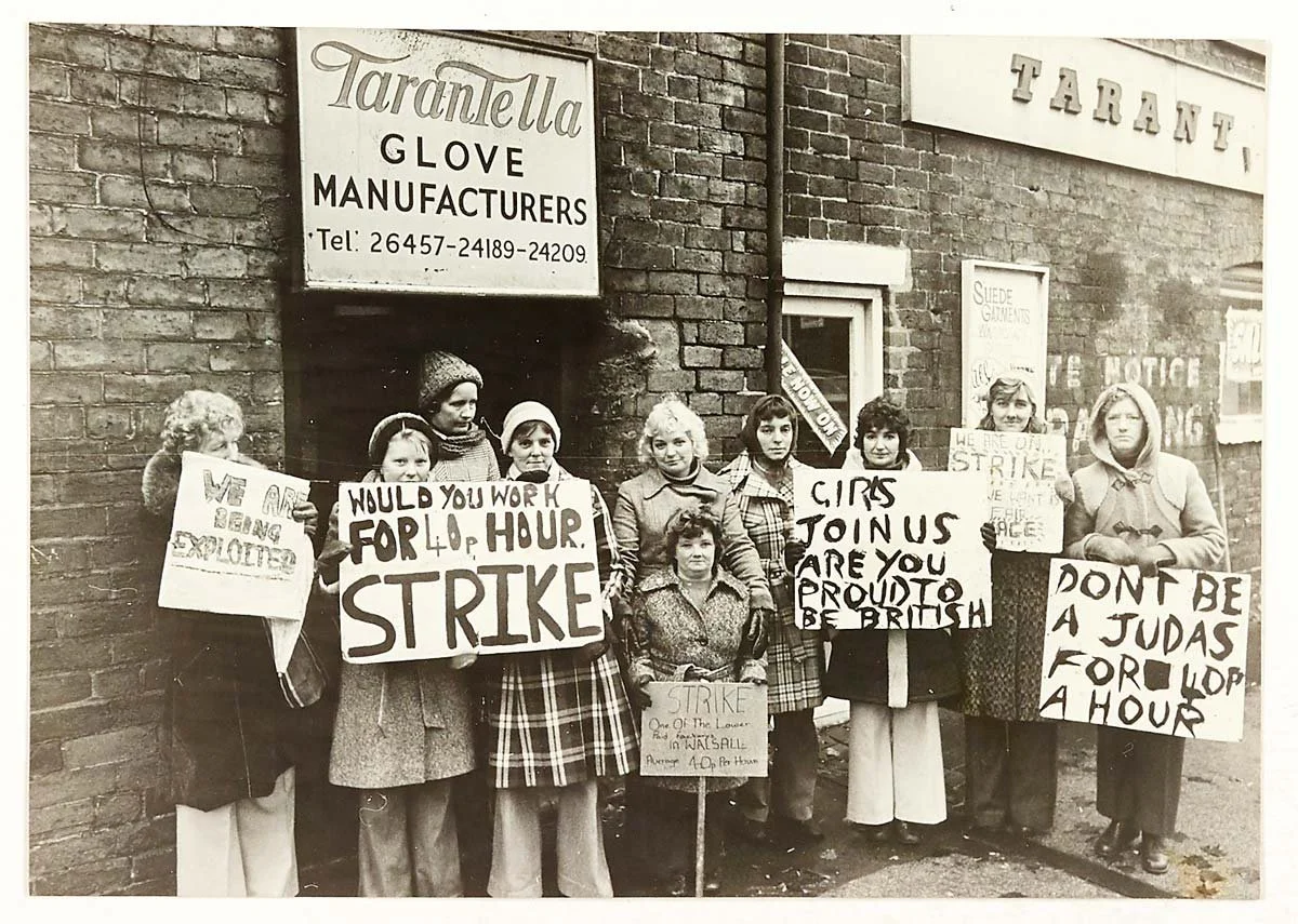 Old newspaper photograph showing women standing outside of a glove manufacturing factory looking serious with placards protesting against low wages.