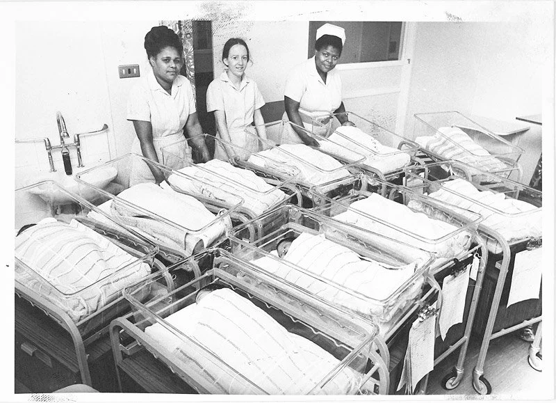 Three young black nurses smiling in a hospital nursery with newborn babies back in the 1960s
