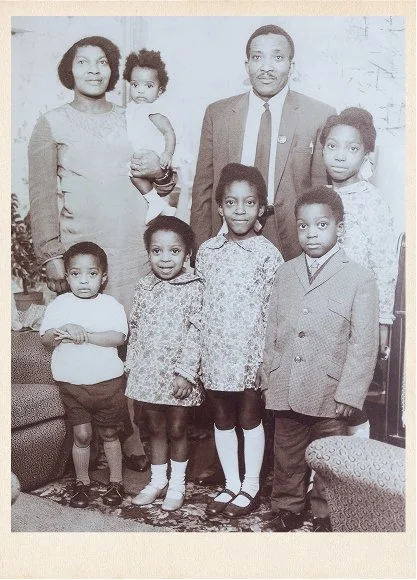 Young jamaican mother and father stood together in a living room with their 6 young children, all dressed in their Sunday best in the 1950s