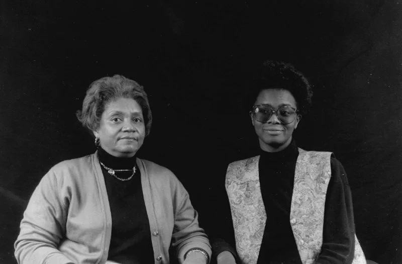 Mature grey haired woman and young black woman sat together before a black backdrop wearing black polo necks underneath smart clothes