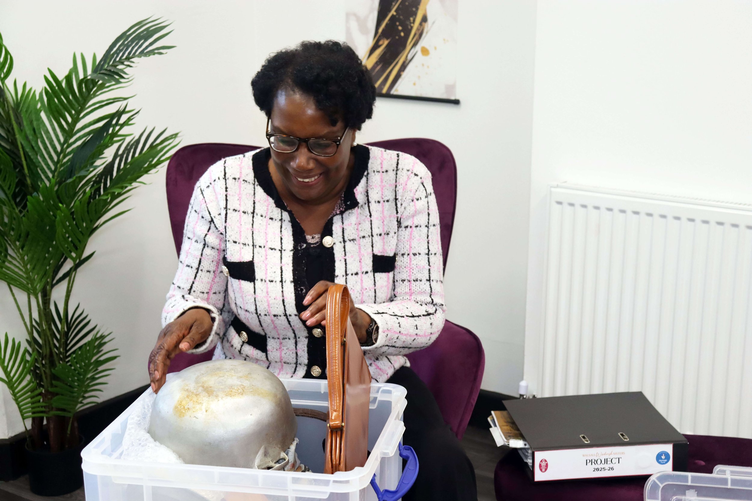A mature woman seating on a comfy purple armchair smiling into a large plastic storage box of assorted heritage items from Caribbean homes including a dutch pot
