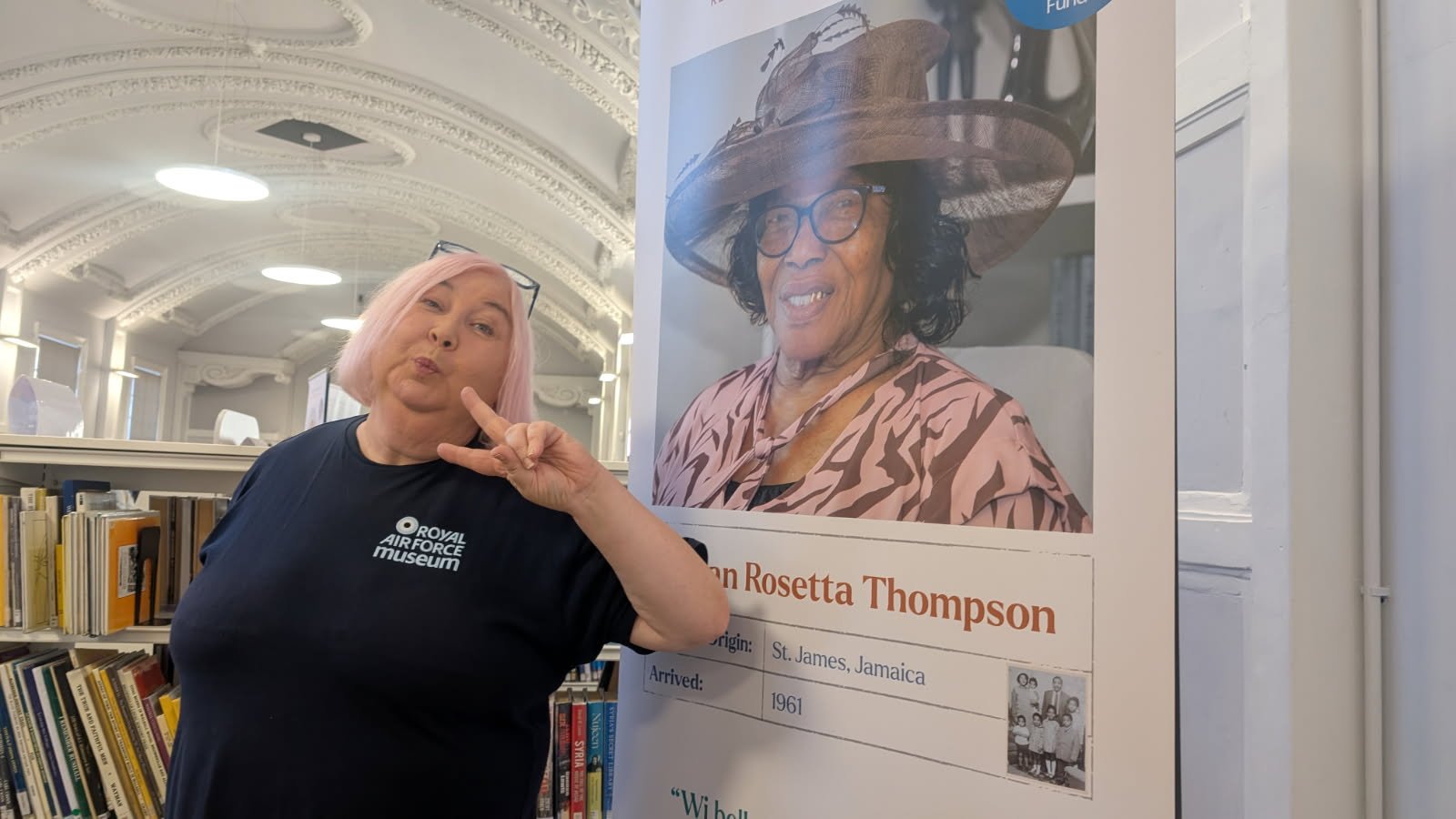 A cheerful woman wearing an RAF Museum t-shirt holding up the peace sign with her fingers playfully stood next to a Walsall Windrush Sister Oral Story Pull-up banner in front of a bookcase