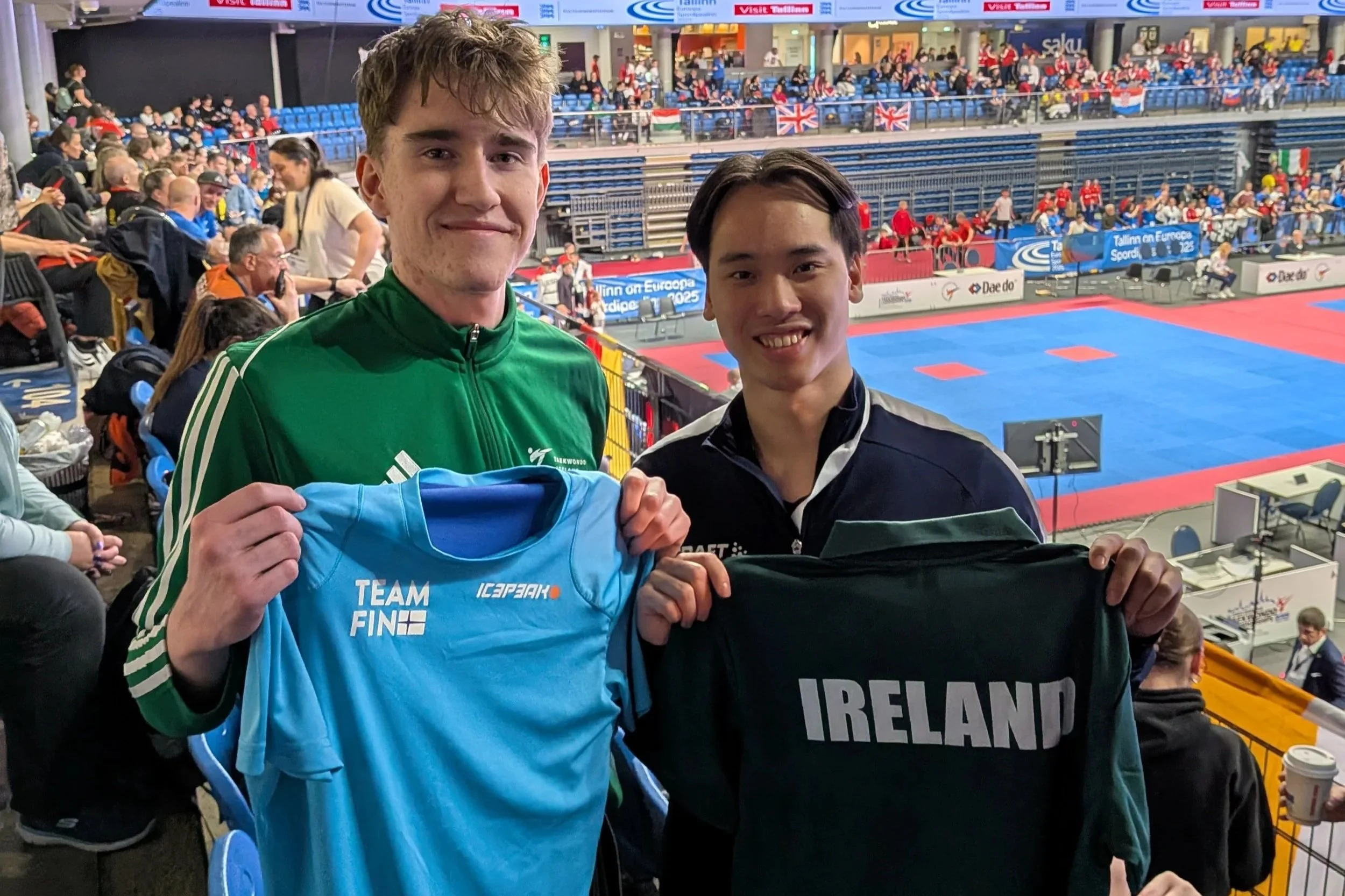 Two young male athletes in a sports arena holding their national team shirts, one with a blue shirt labeled 'TEAM FIN' and the other with a black shirt labeled 'IRELAND'. The arena features a martial arts or taekwondo competition setup with spectator