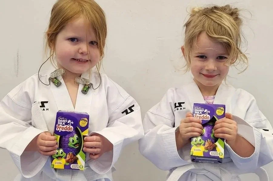 Two young girls dressed in white martial arts uniforms holding cartons of Cadbury Dairy Milk Freddo chocolate, standing barefoot on a blue mat against a plain wall.