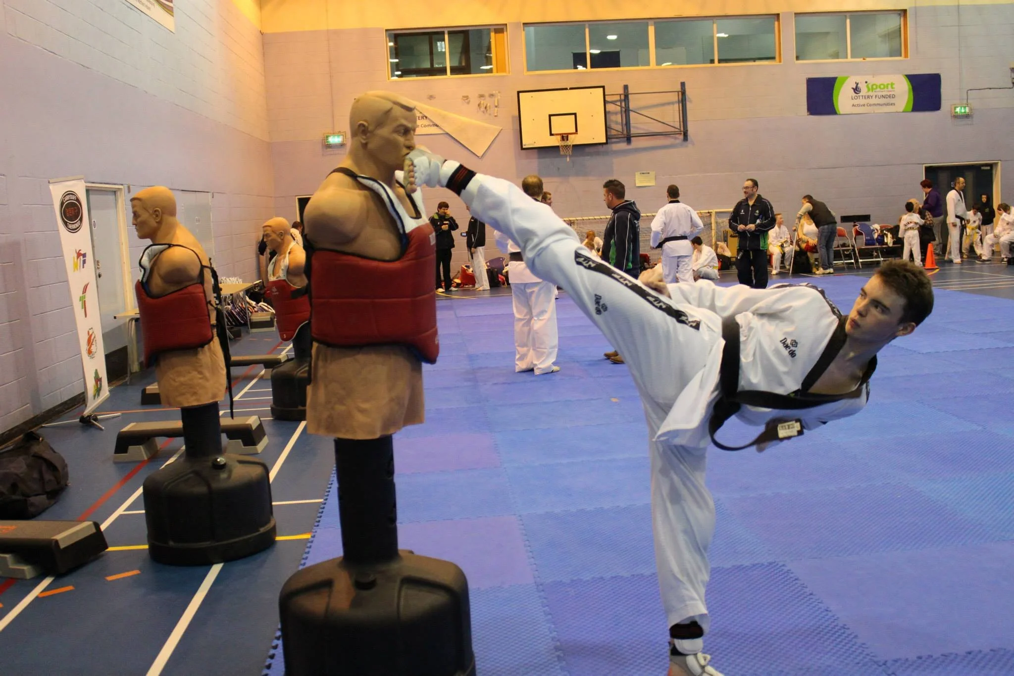 A martial artist in a white uniform executing a high kick towards a punching dummy at a martial arts training session in a gymnasium.