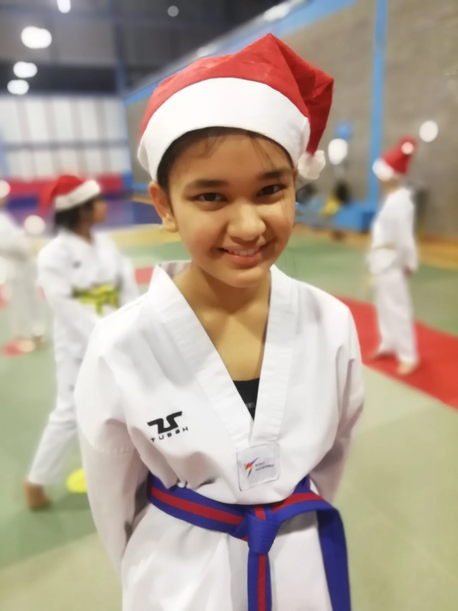 Young martial artist wearing a Santa hat and a white taekwondo dobok with a blue and red belt, smiling inside a gym with other children practicing martial arts.