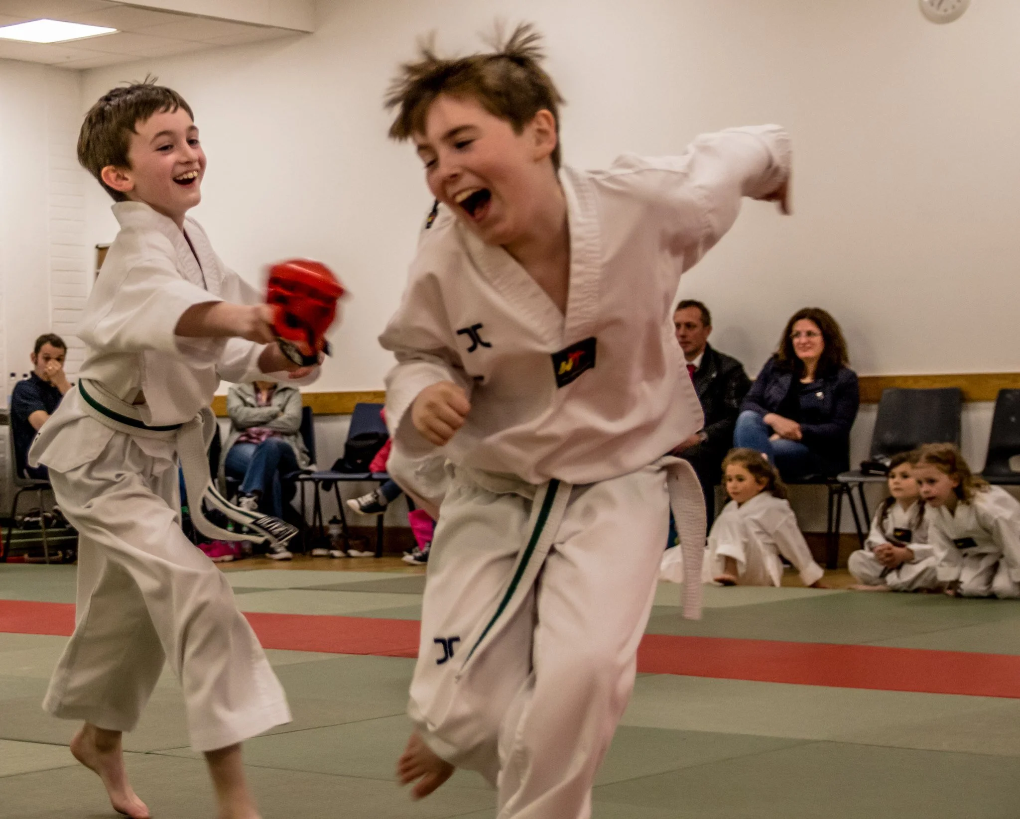 Two young boys in martial arts uniforms playing and smiling during a martial arts class or competition, with an audience of children and adults sitting in the background.