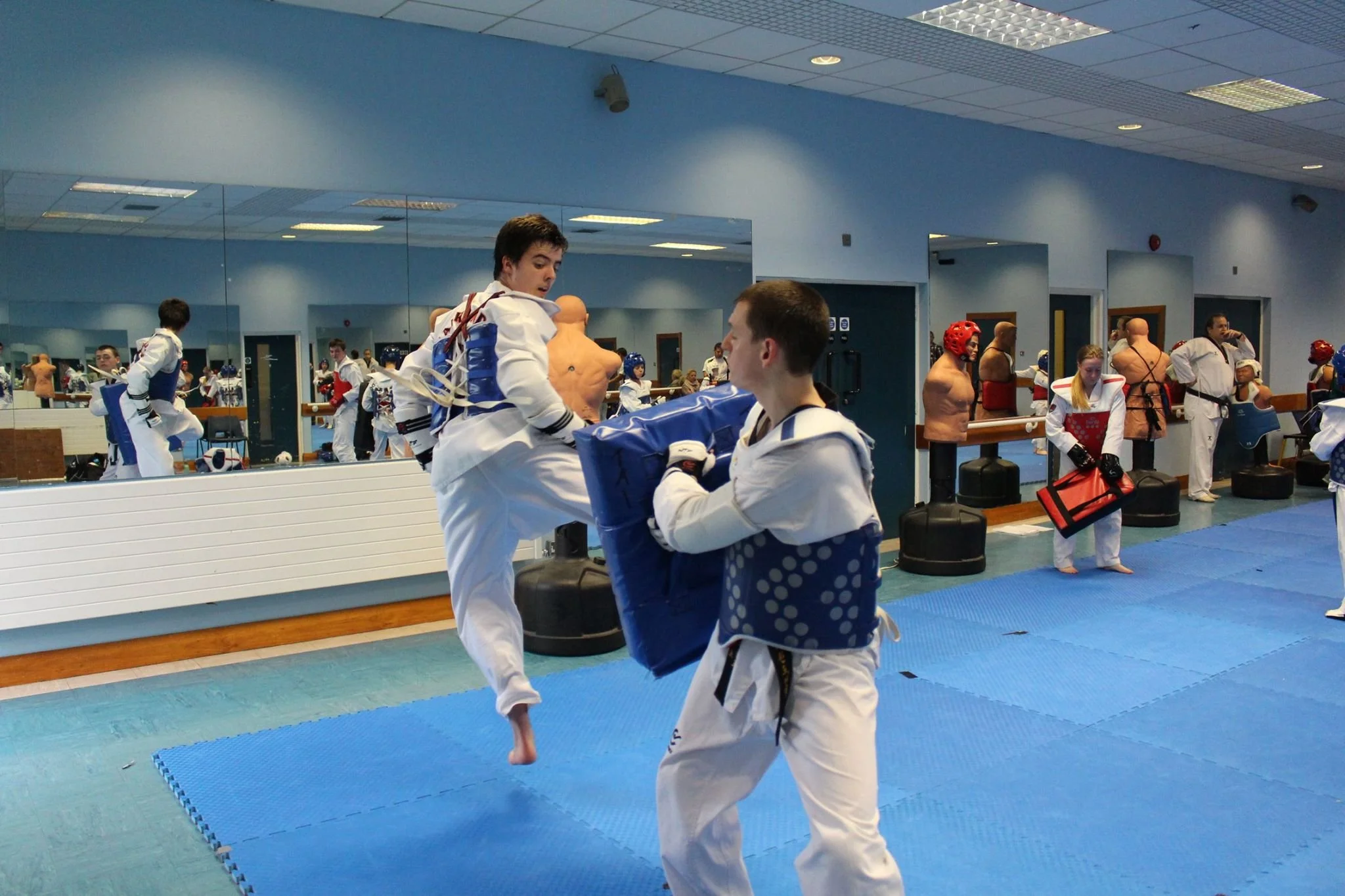 Martial arts students practicing sparring techniques in a dojo with mirrors, padded mats, and training equipment.