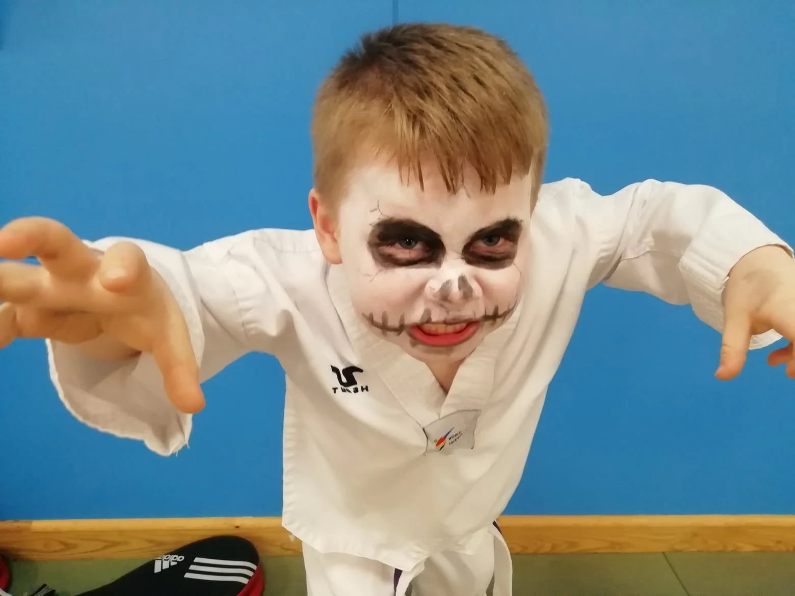A young boy with face paint resembling a skull, wearing a martial arts uniform, leaning forward with hands extended toward the camera, against a blue wall and wooden floor.