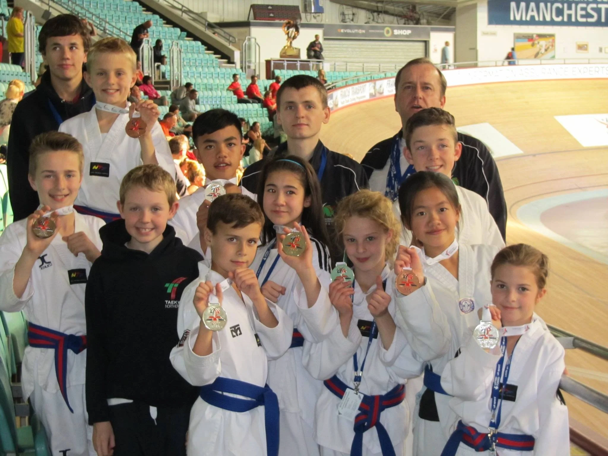 A group of young martial artists in uniform, smiling and showing medals at a sports venue, with a coach or instructor among them and an indoor stadium in the background.