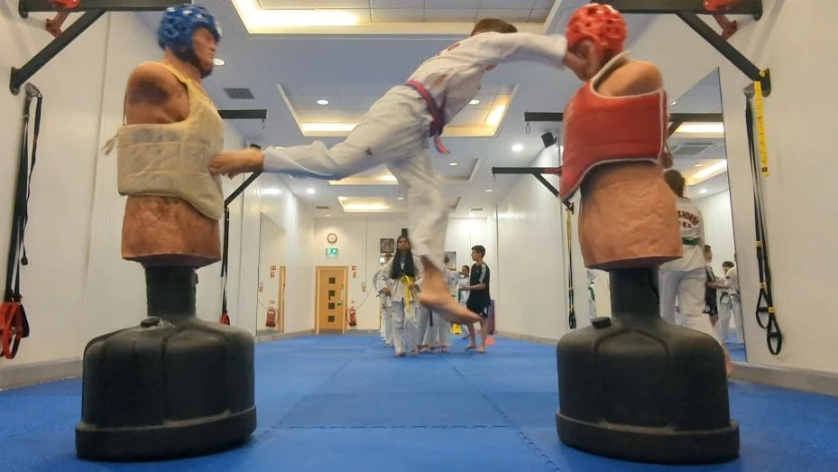 Martial arts dojang with children practicing and sparring, with two punching bags in the foreground.