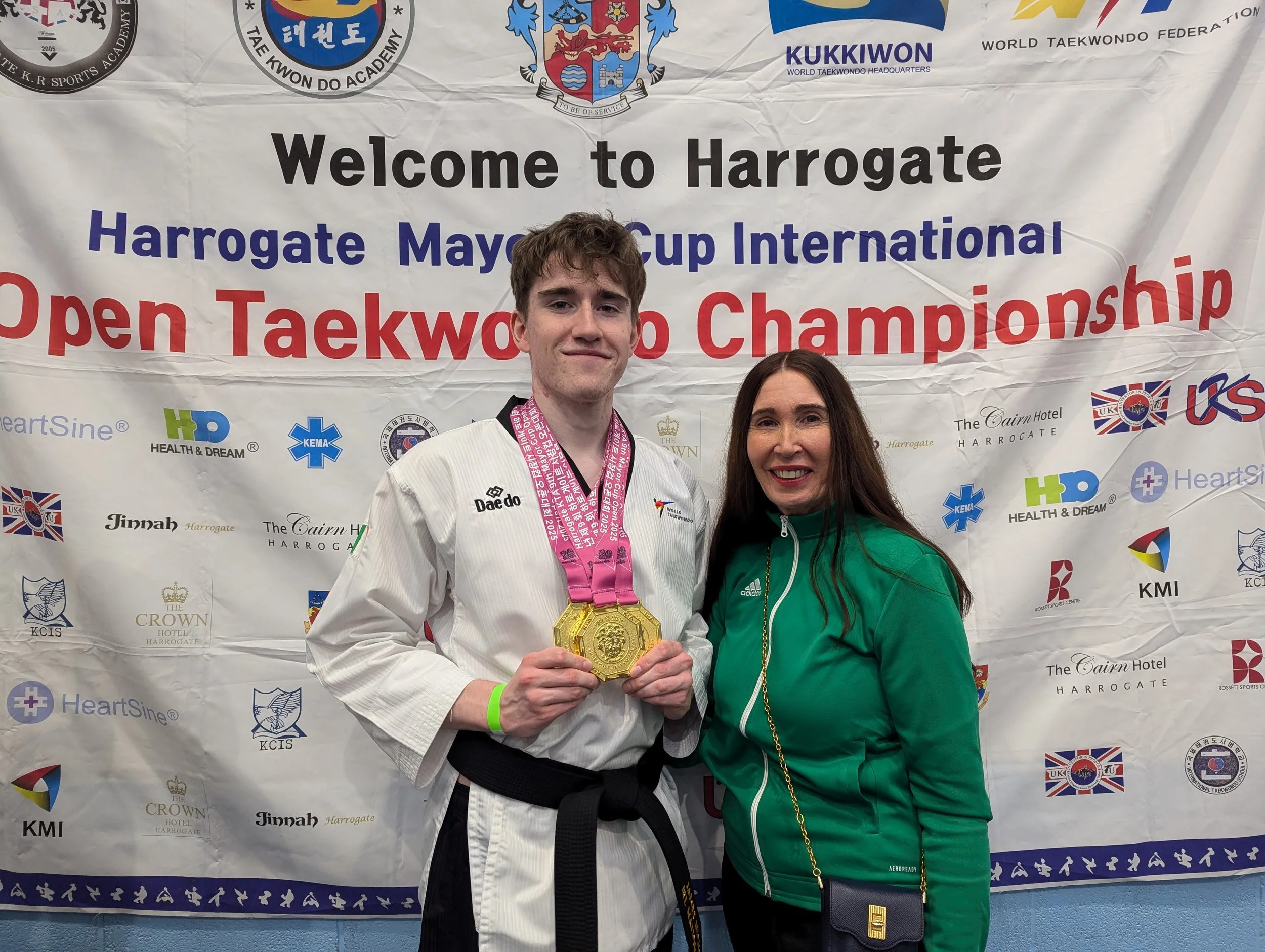 A young man in a white martial arts uniform with a black belt, holding a gold medal, standing next to a woman in a green jacket, both smiling for a photo in front of a banner for the Harrogate Mayors Cup International Open Taekwondo Championship.