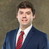 Portrait of a young man in a navy suit, white shirt, and red striped tie, smiling against a neutral background.