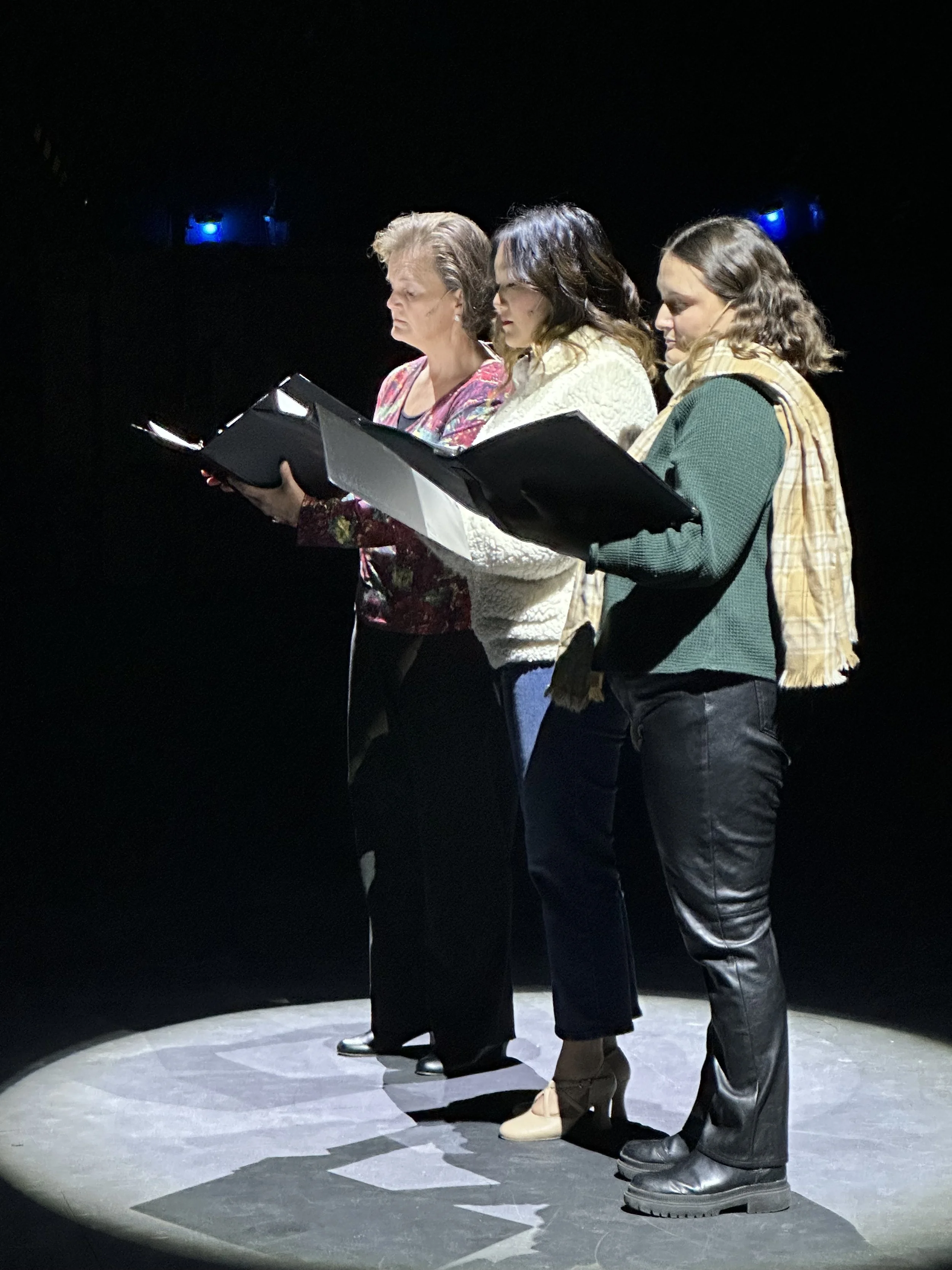 Three women hold scripts or folders, standing under a spotlight on a dark stage, appearing focused during a rehearsal or performance.