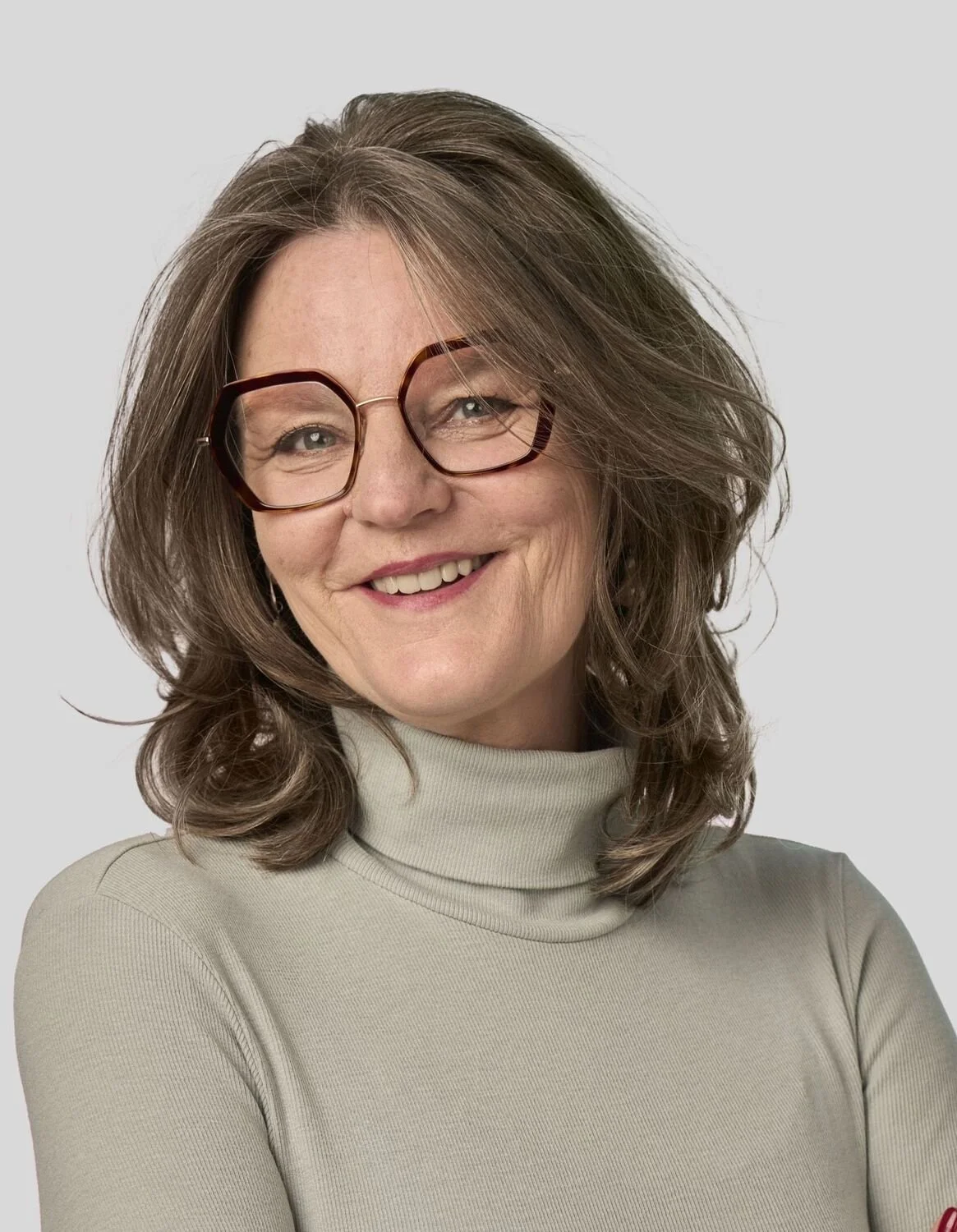 Eine lächelnde Frau mit braunen, gewellten Haaren trägt große Brillen und ein beige Turtleneck.