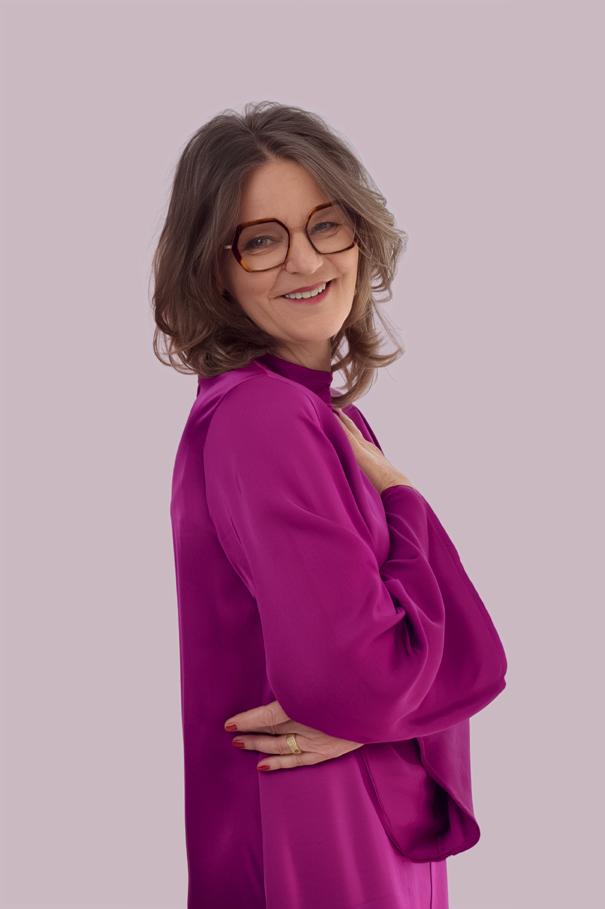 Eine lächelnde Frau mit Brille, in einer magentafarbenen Bluse und Kleid, posiert vor einem hellvioletten Hintergrund.