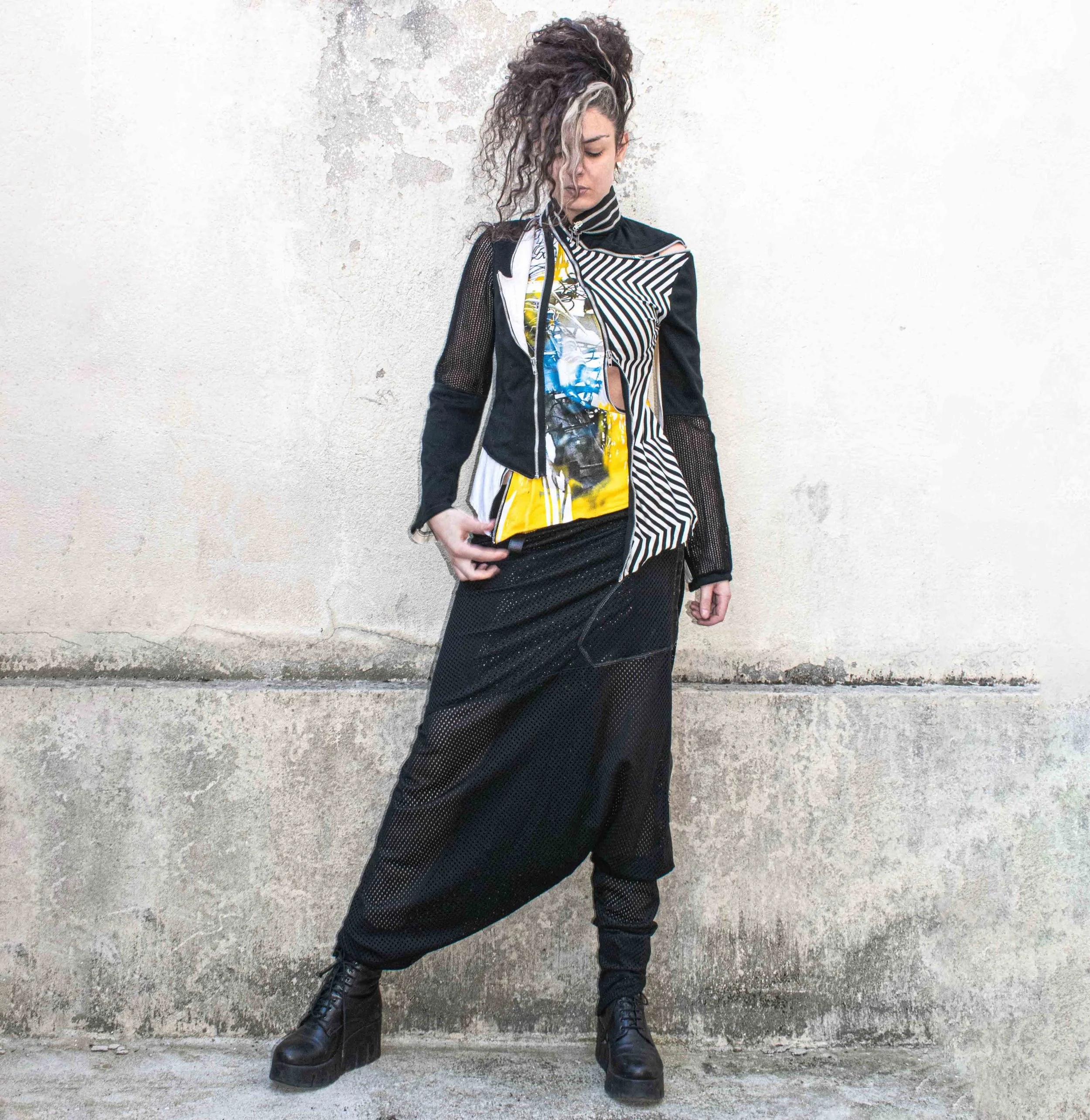 A woman with curly hair and multiple colored streaks, wearing eclectic black and patterned clothing, standing against a concrete wall.