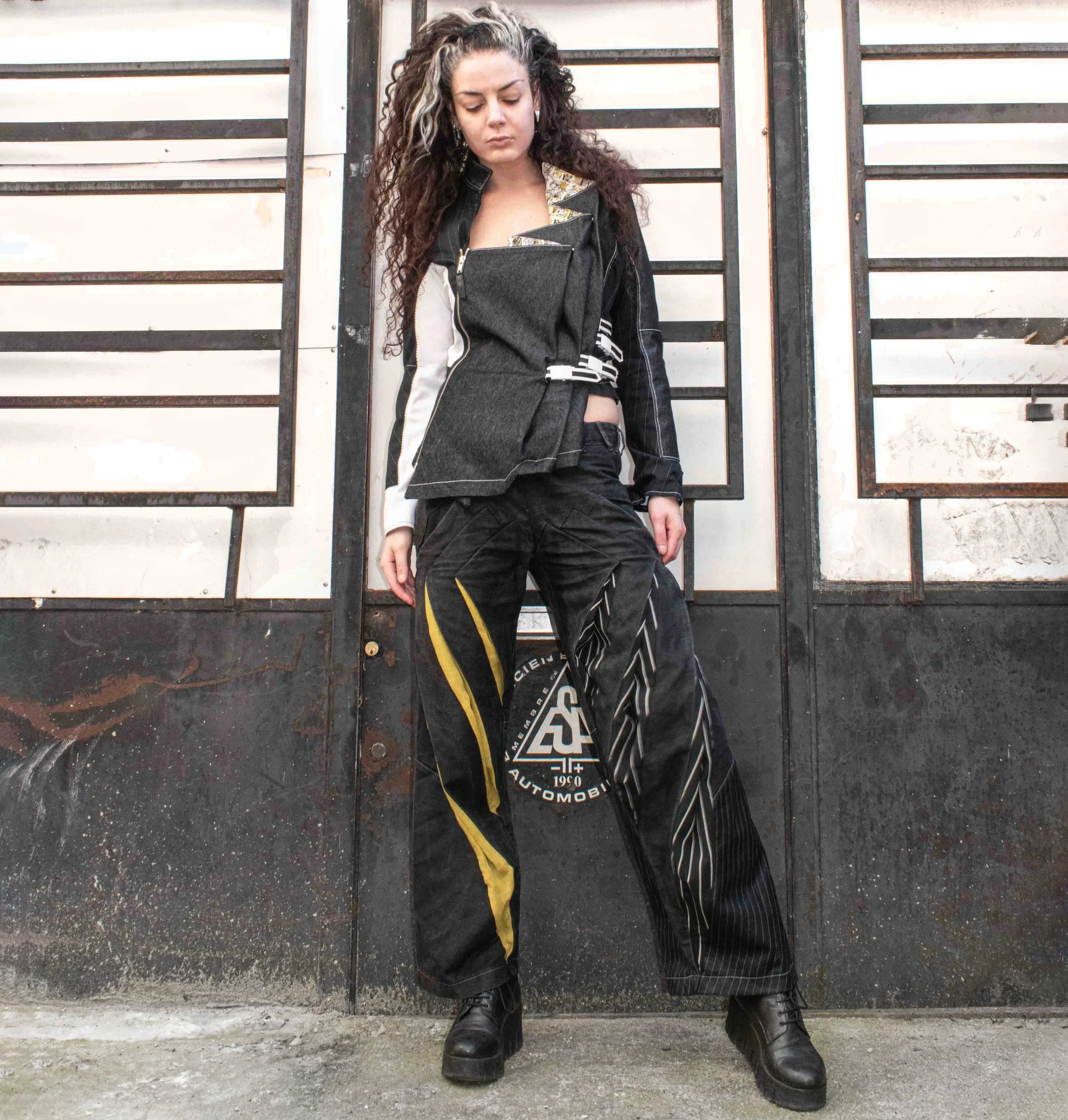 Young woman with curly hair in edgy streetwear, standing outdoors in front of a black and white metal fence with a circular logo on the wall behind her.