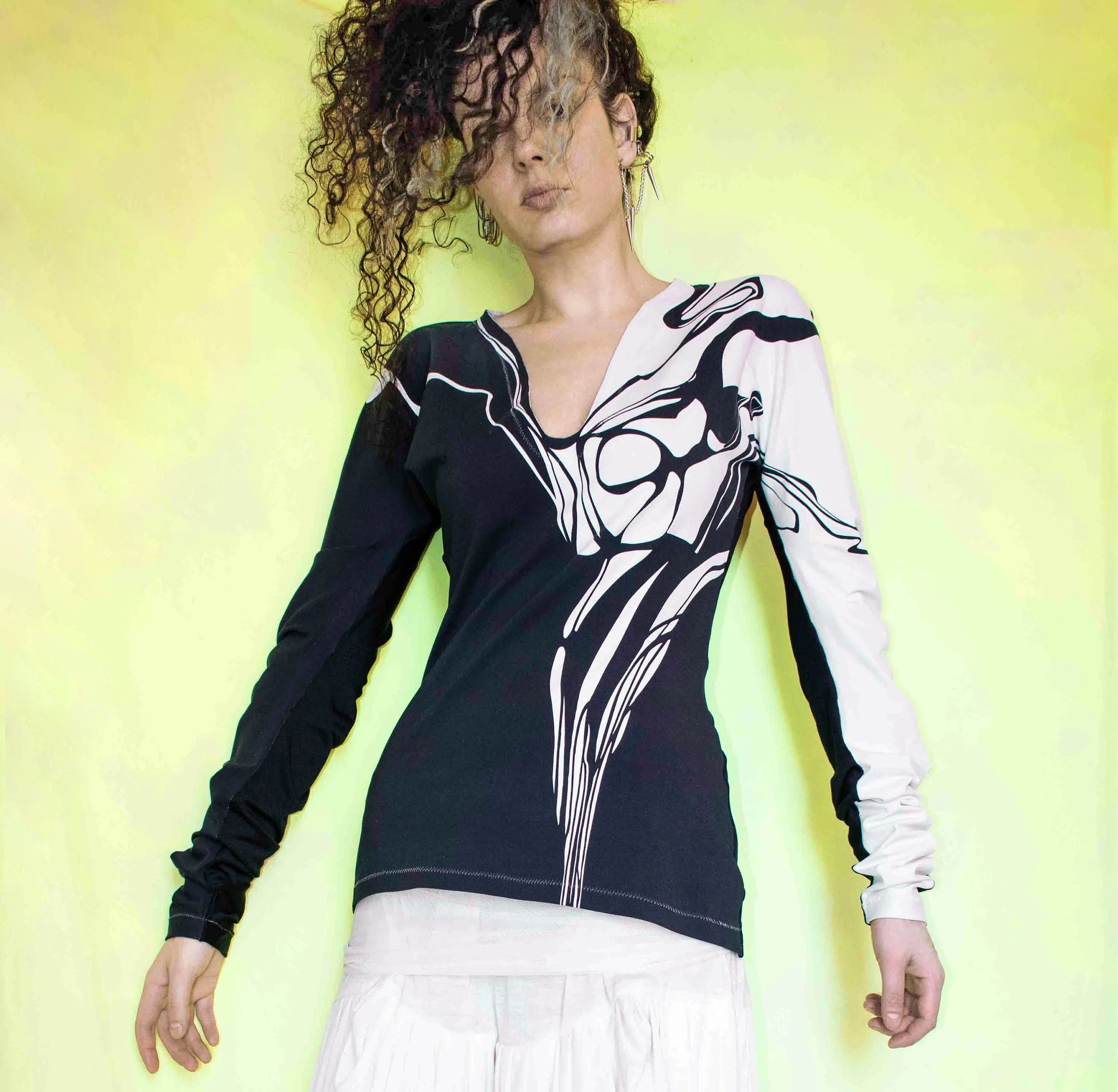 A woman with curly hair styled in an asymmetrical fashion, wearing a black and white long-sleeve top with abstract lines, and a white skirt, standing against a light yellow background.