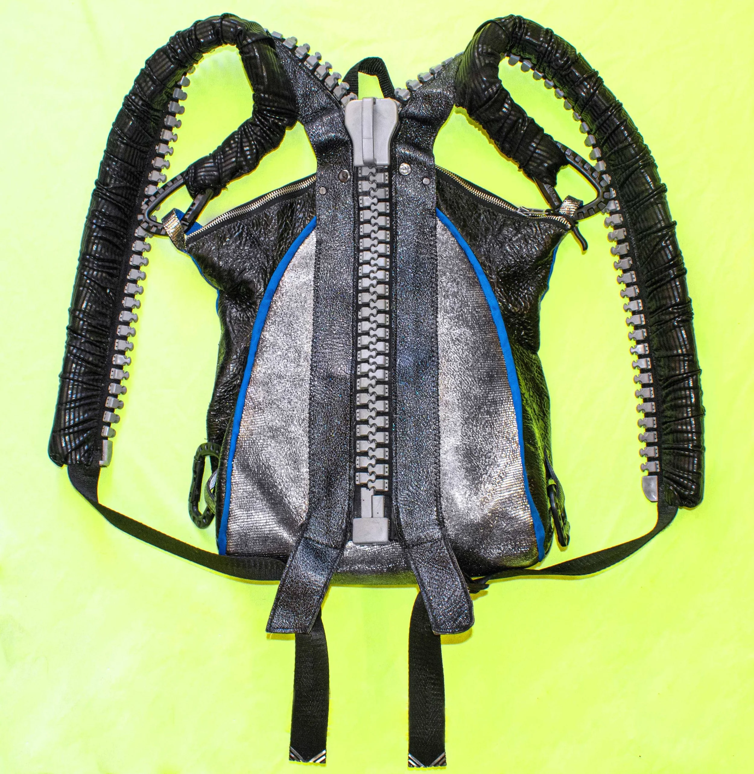 A black and silver backpack with multiple zippered compartments and thick, adjustable shoulder straps, placed on a bright yellow background.