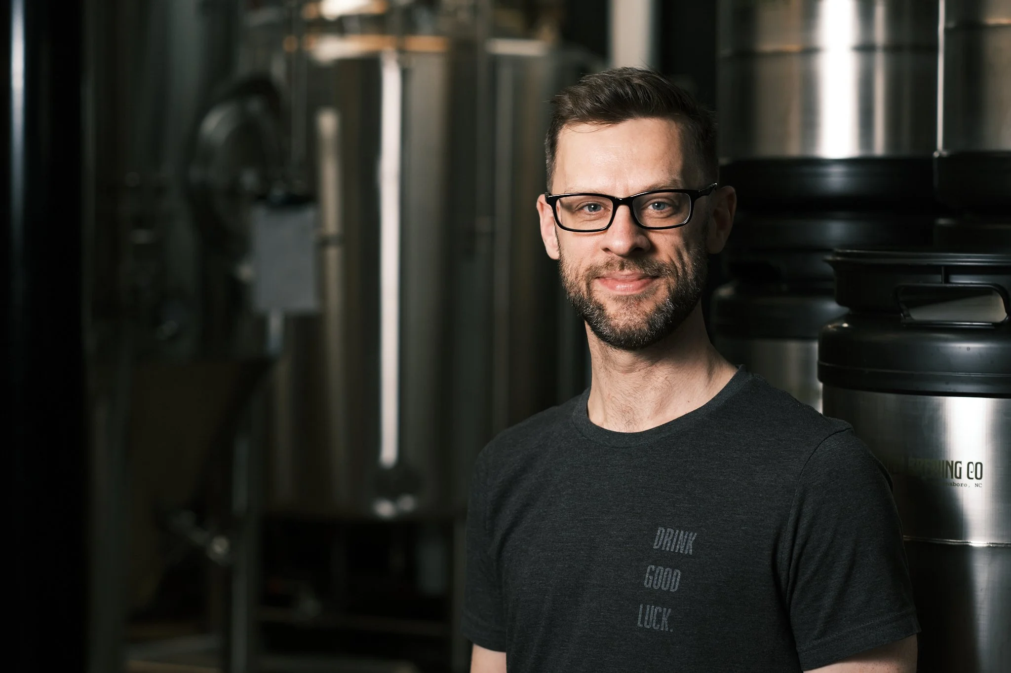 A man with glasses and a beard standing in front of large metal fermentation tanks in a brewery, wearing a black T-shirt that says "DRINK GOOD LUCK."