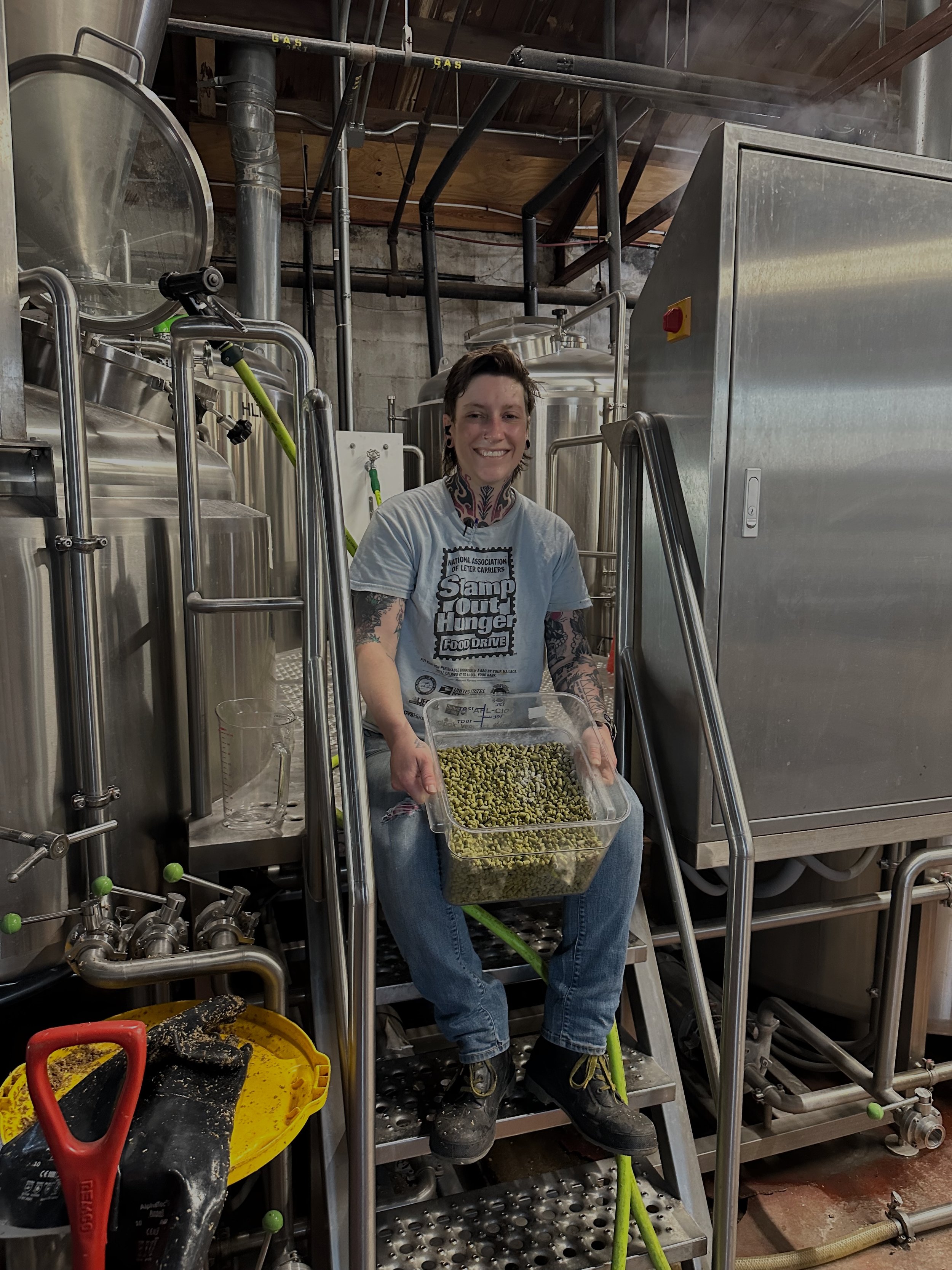 A woman with tattoos on her arms and neck, smiling, sitting on a metal platform in a brewery, holding a clear plastic container filled with green hops, surrounded by brewing equipment.