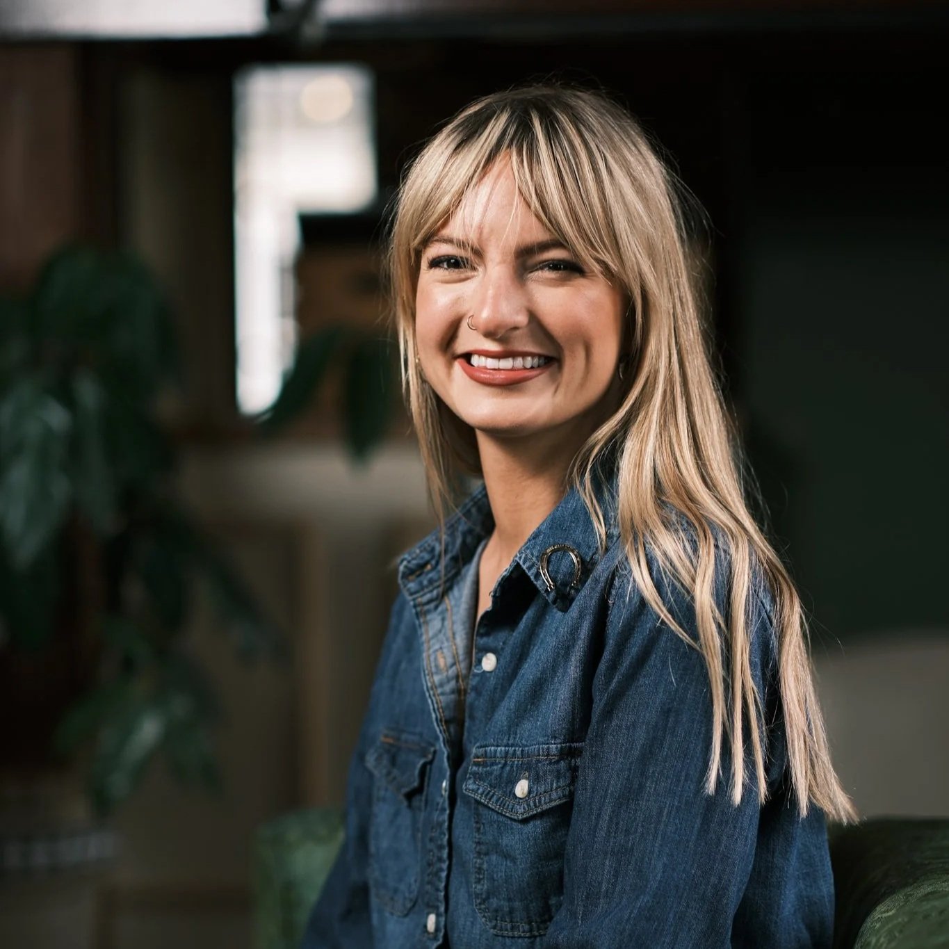 A smiling woman with blonde hair and a nose ring, wearing a denim shirt, sitting indoors with a dark background and plants.