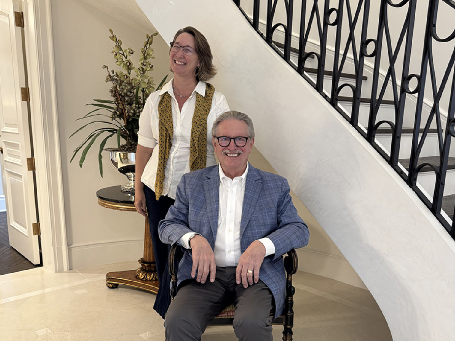 A smiling man in a blue checkered blazer seated on a vintage wooden chair, with a woman standing behind him, both near a staircase with black railing, and a table with a silver plant pot and a large plant next to a white wall.