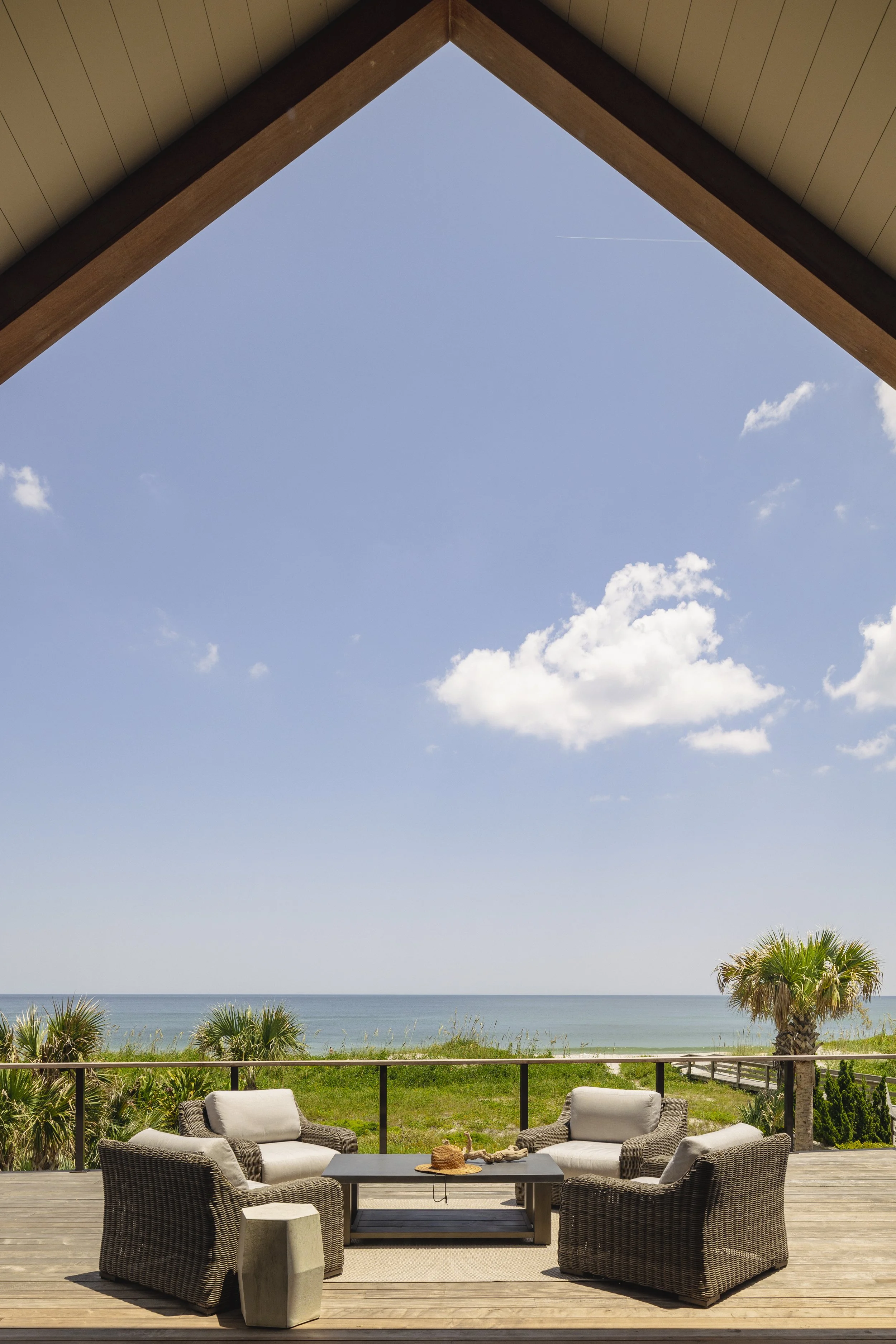 A sunny outdoor patio with wicker chairs and a table, overlooking a beach with palm trees and the ocean under a blue sky with scattered clouds.