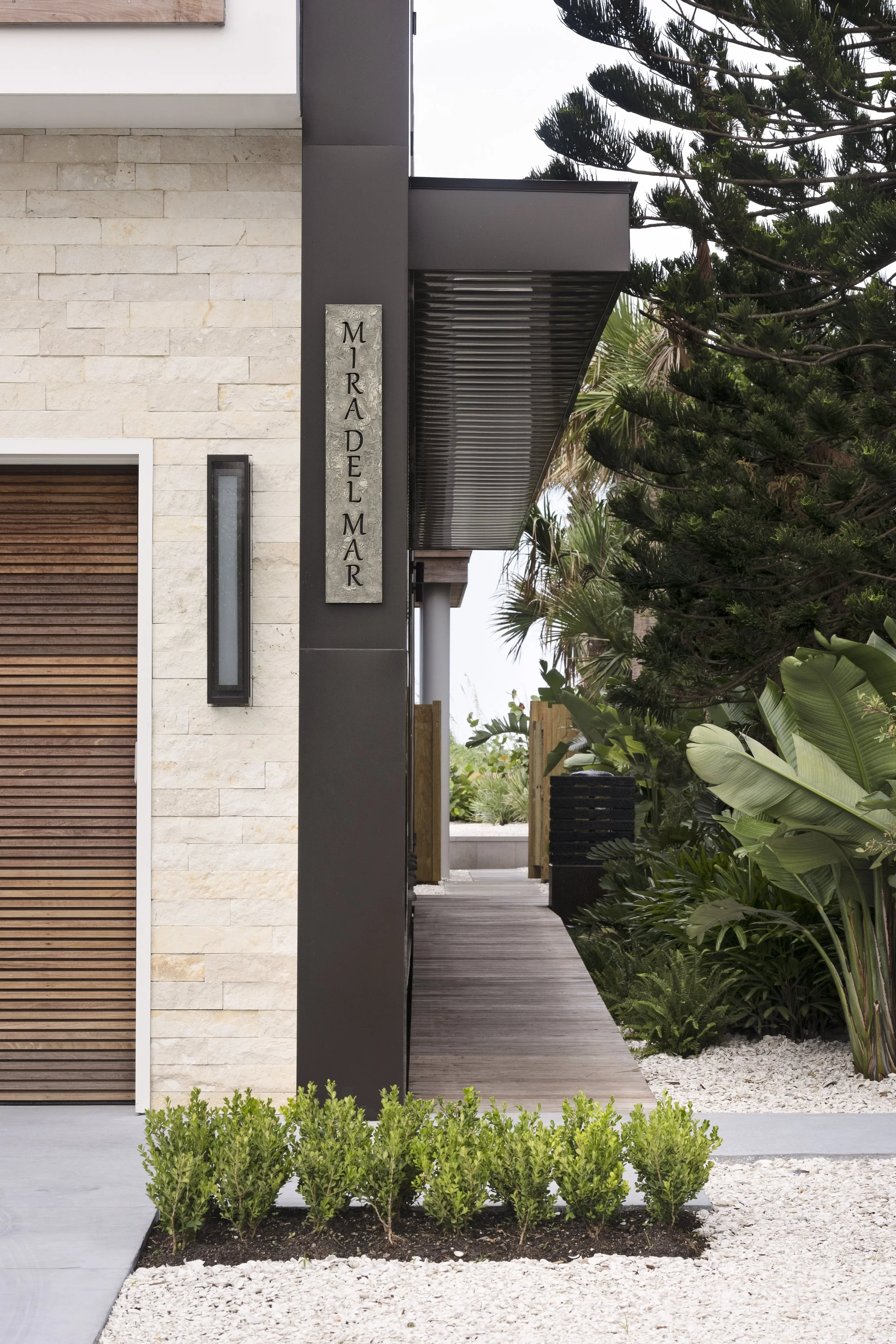 Modern house exterior with a stone and dark metal facade, lush green plants, and a wooden walkway leading to an outdoor area.