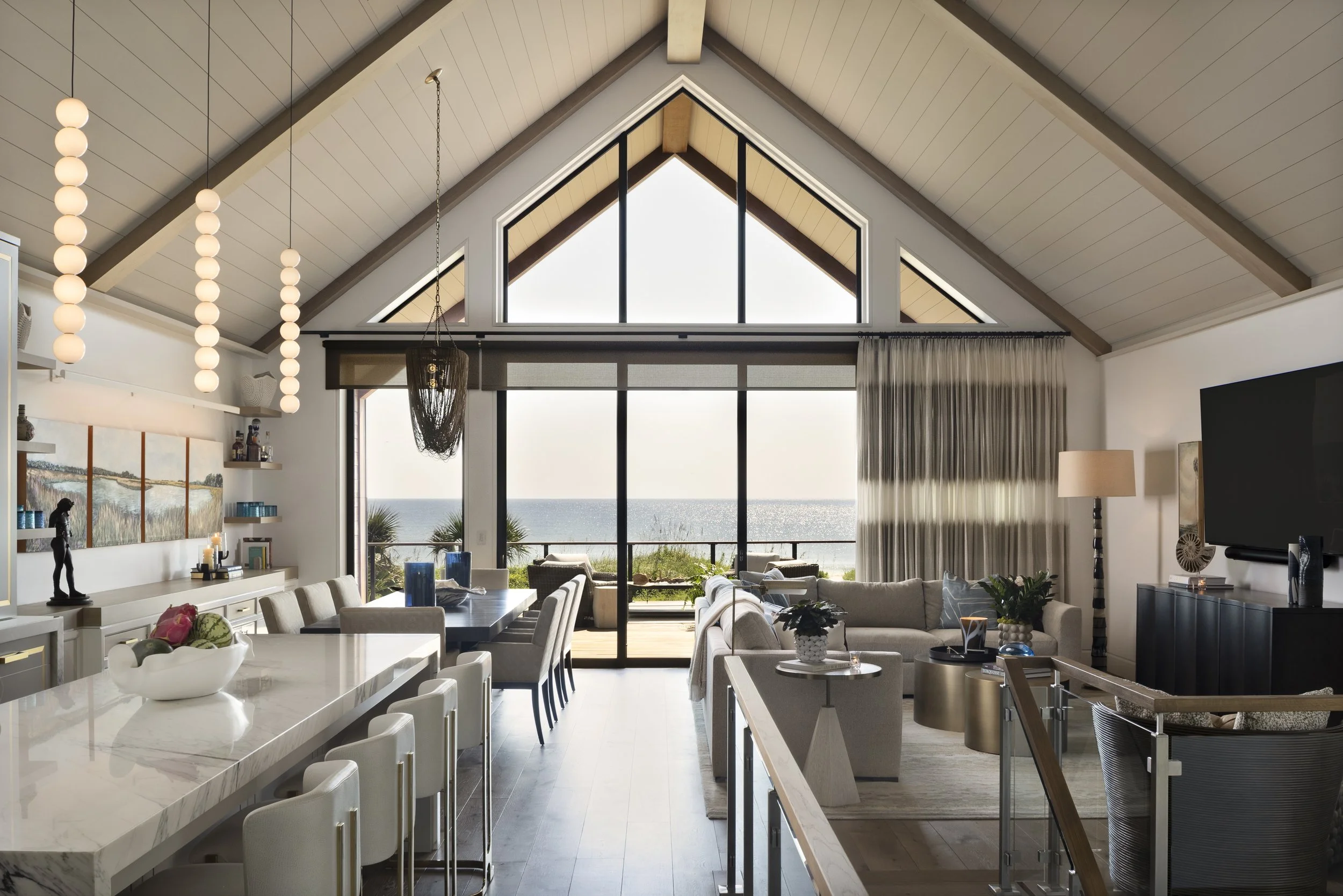 Modern living room with large glass windows and a view of the ocean, featuring a high vaulted ceiling with wooden beams, a dining area with a marble-topped table, and a cozy seating area with sofas and a flat-screen TV.