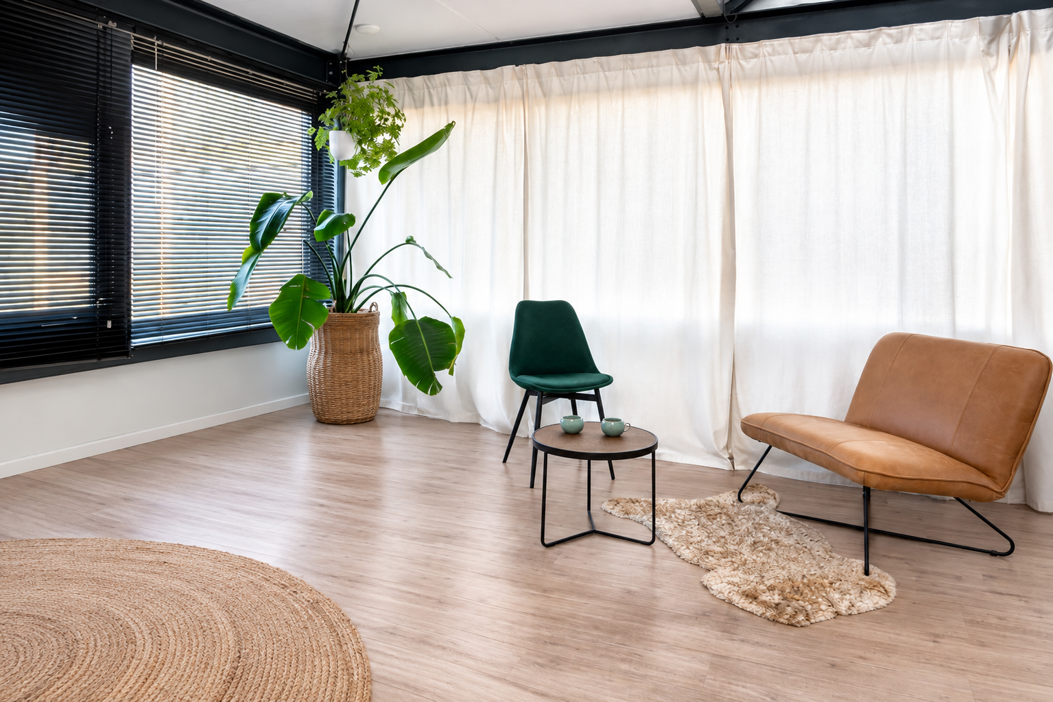 Een woonkamer met grote ramen, zwarte jaloezieën, een grote groene plant in een mand, een fauteuil, een bank, een ronde salontafel met twee koppen, een klein tapijt en een stoffen gordijn.