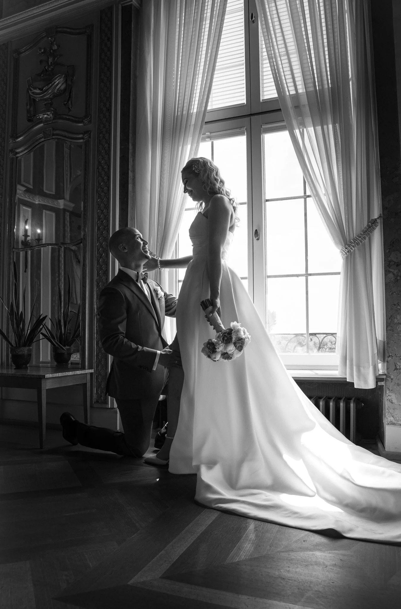 Braut und Bräutigam bei ihrer Hochzeit, braut steht vor braut, während der Bräutigam kniet und sie ansieht, im Raum mit großen Fenstern und eleganter Inneneinrichtung. Fotografiert von Kamisight Photography, in Hessen.